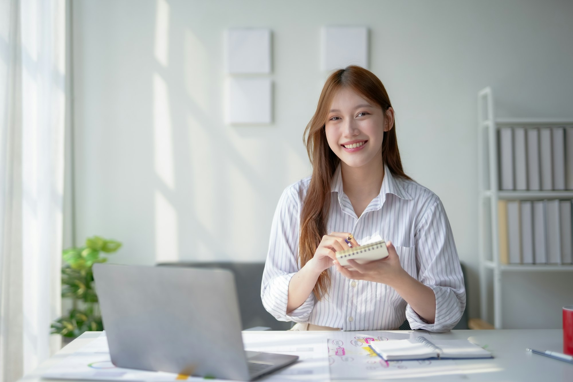 Smiling woman working from home, holding notepad, and sitting at a desk with laptop